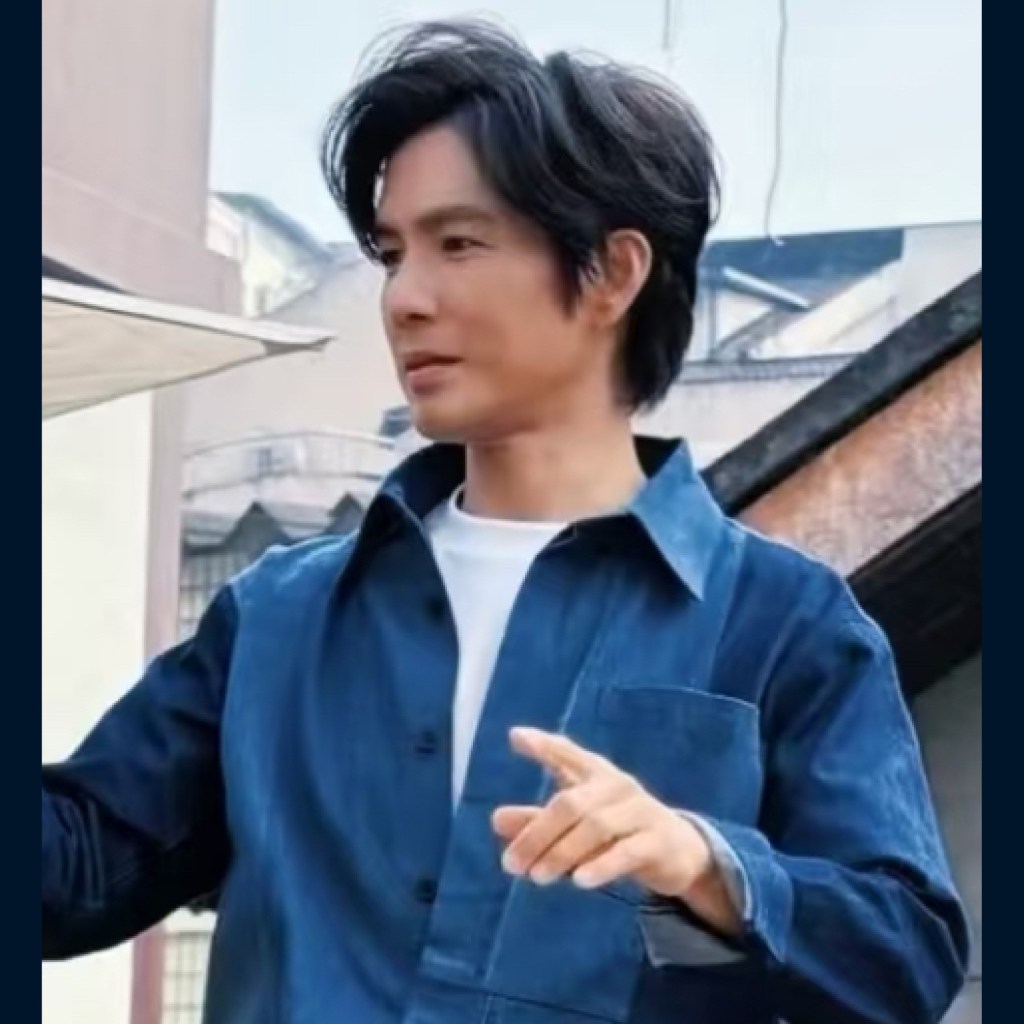 A man wearing a blue denim shirt gestures thoughtfully while standing outdoors. He appears to be engaged in conversation.